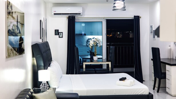 A modern hotel room with a bed, a desk, a chair, a small table, and a balcony with a view. - Kandy Haven - Studio Type Unit (Angeles City)