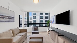 Een moderne woonkamer met een bank, poef, televisie en een groot raam met uitzicht over de stad.