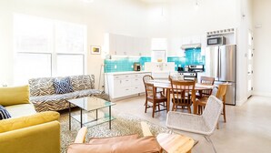 Uma cozinha moderna com armários brancos, uma sala de jantar com mesa de vidro e uma sala de estar com sofá e poltronas.