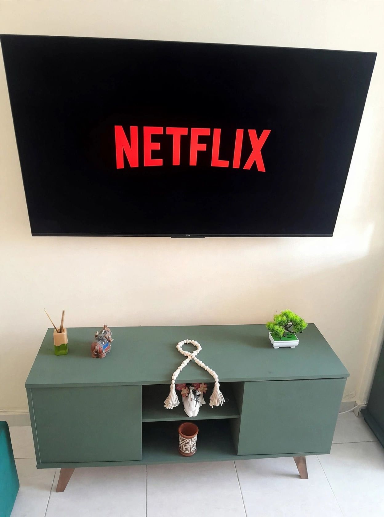 Ein Fernseher mit Netflix-Logo auf dem Bildschirm, eine grüne Unterhaltungsecke mit Dekorationselementen und ein kleiner Topfpflanze.