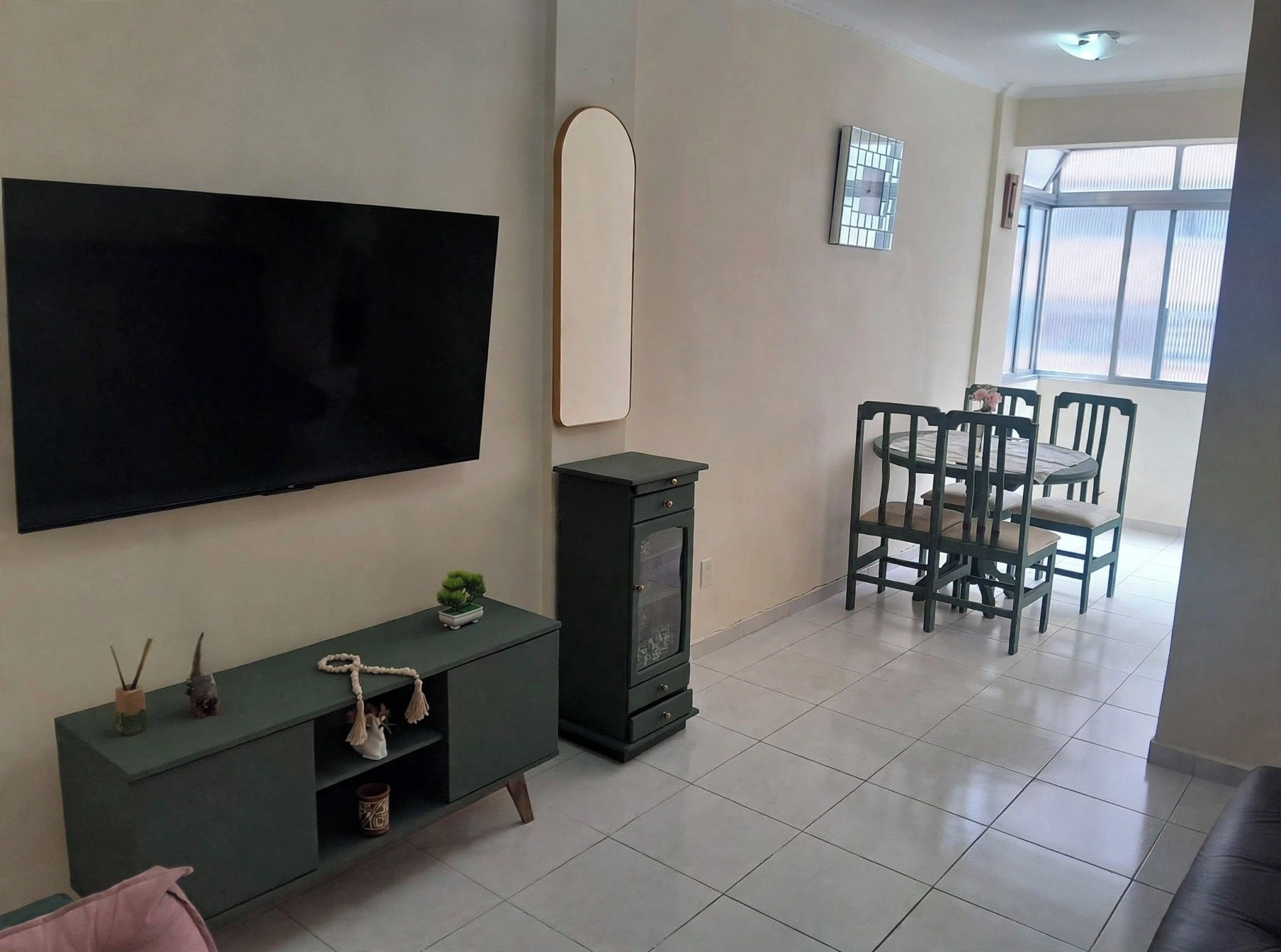 A living room with a flat-screen TV, a dark-colored entertainment unit, a dining area with a table and chairs, and a window with blinds.