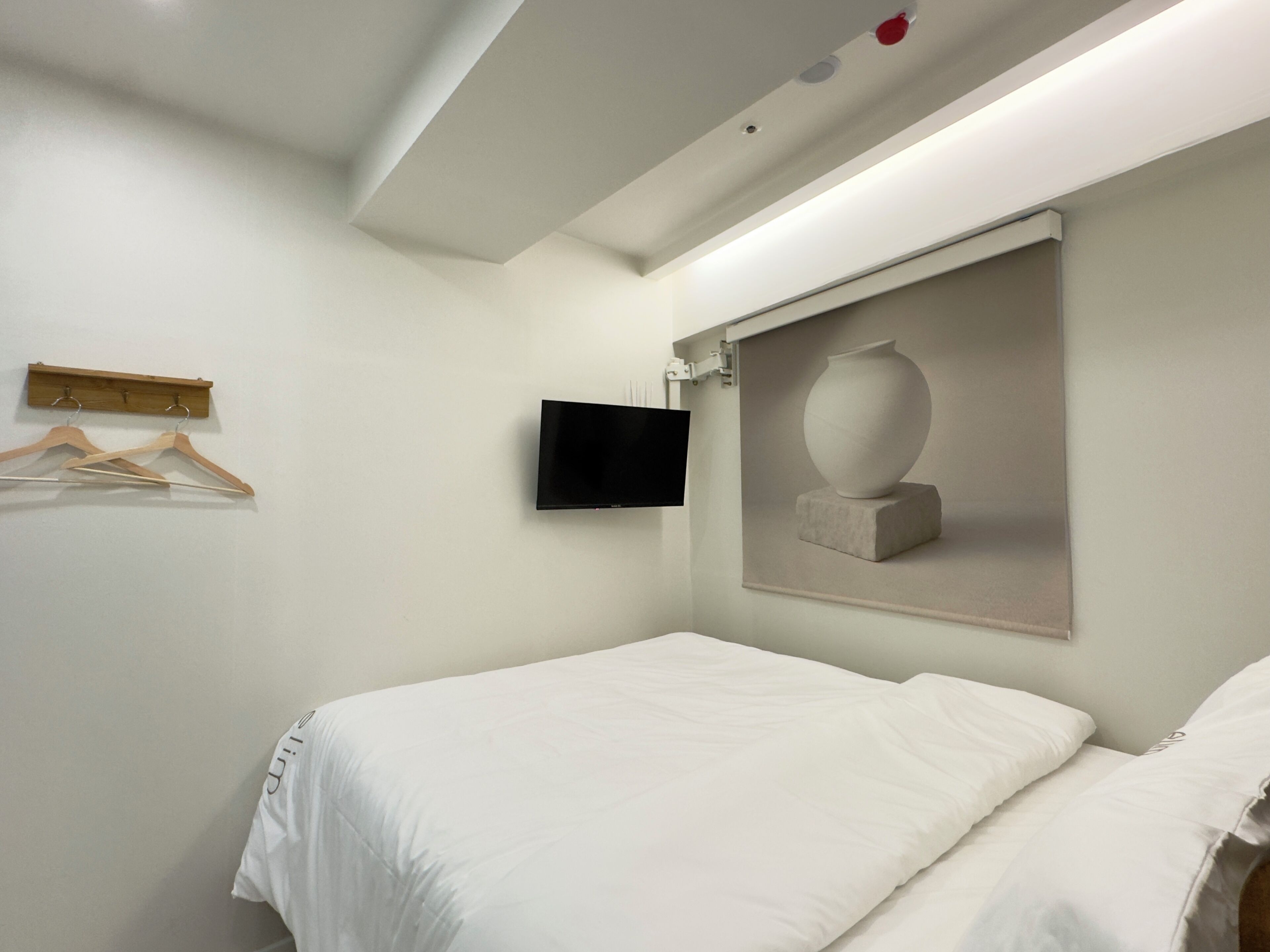 Una habitación de hotel con una cama, un televisor montado, un perchero de madera y una obra de arte enmarcada en la pared.