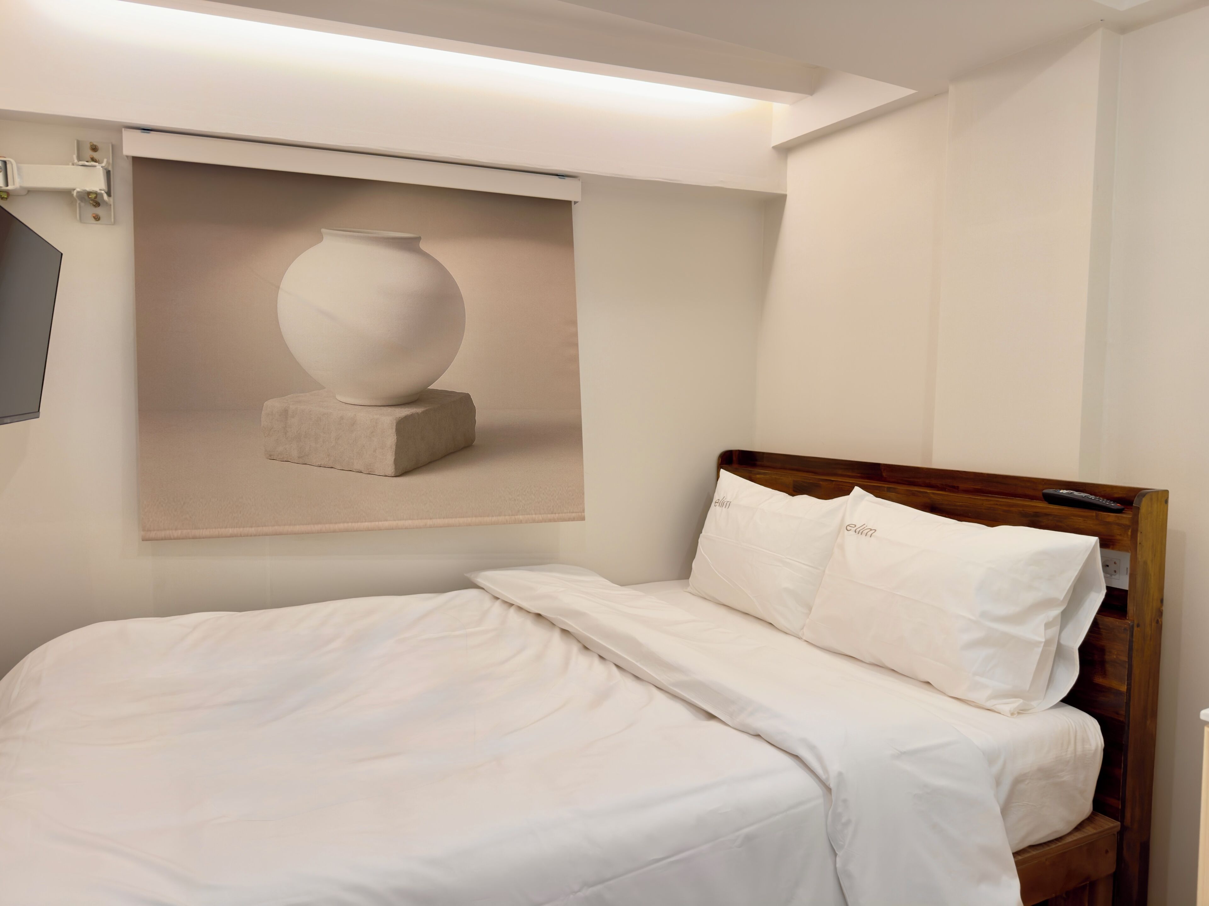 A neatly made bed with white linens, a wooden headboard, and a remote control on the right side.