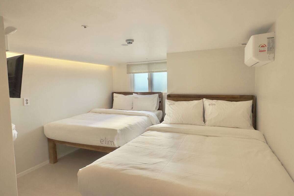 A hotel room with two beds, a window with blinds, a mounted air conditioner, and a ceiling-mounted smoke detector.
