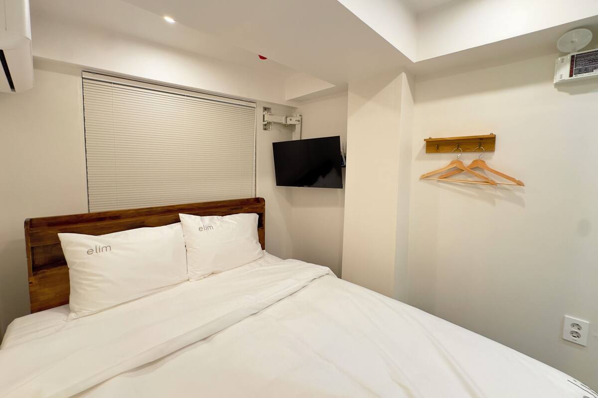 A bedroom with a wooden headboard, white bedding, a mounted TV, a window with blinds, and a coat rack with hangers.