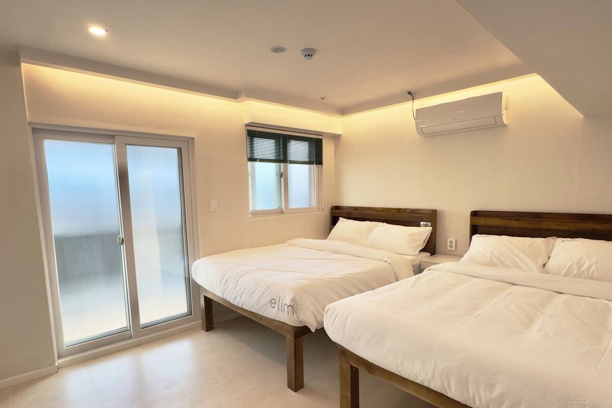 A bedroom with two beds, a sliding glass door, a window with blinds, and an air conditioning unit.