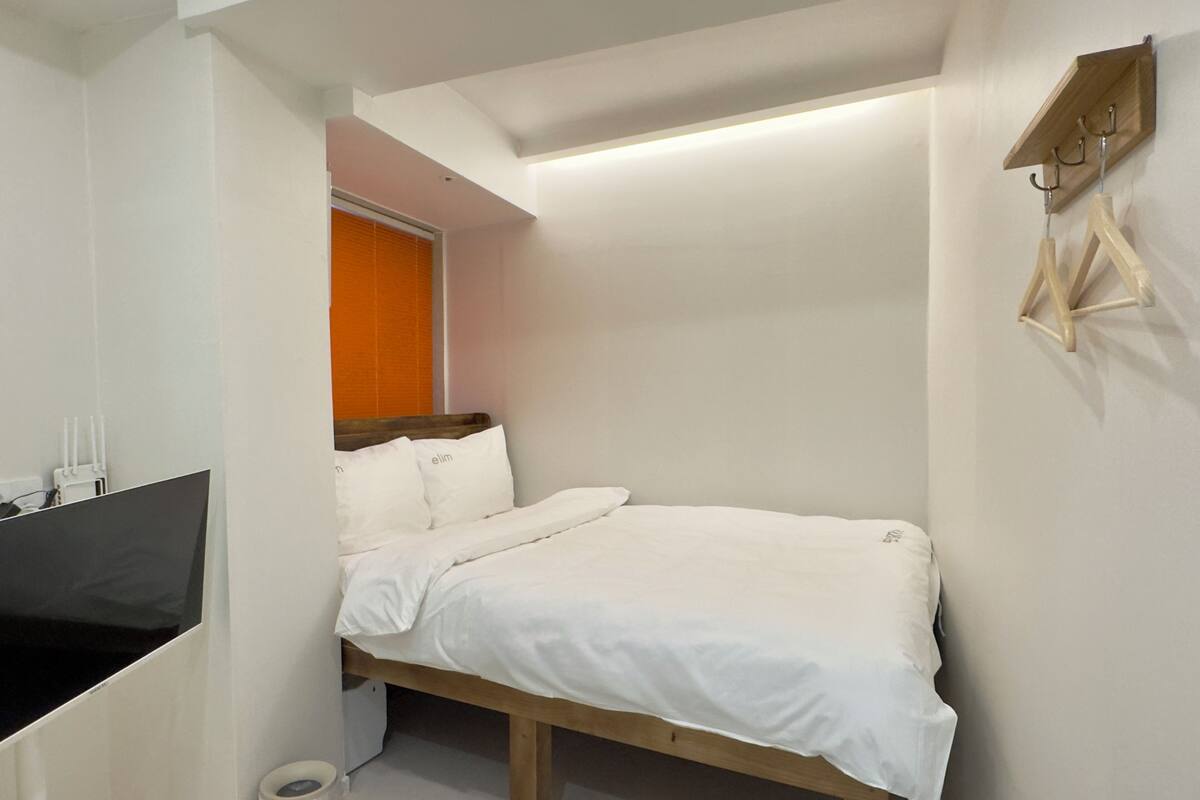 A small, compact bedroom with a single bed, a TV, and a wooden shelf with hooks.
