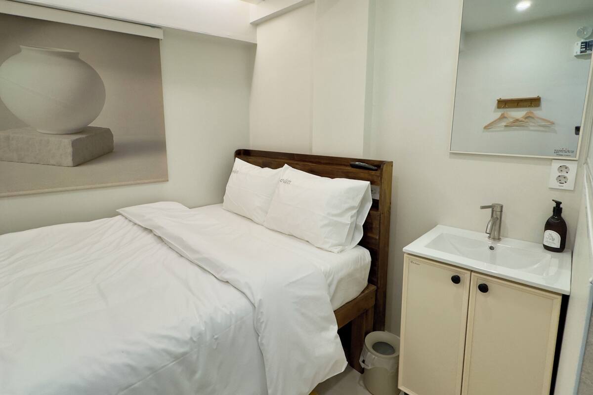 A hotel room with a bed, a wooden headboard, a white bedside table, a mirror, and a small sink.