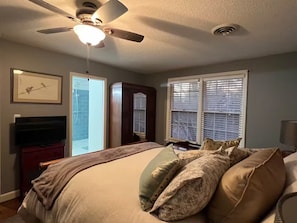 Une chambre à coucher comprenant un lit, un ventilateur de plafond, une télévision, une armoire et une fenêtre avec des stores.