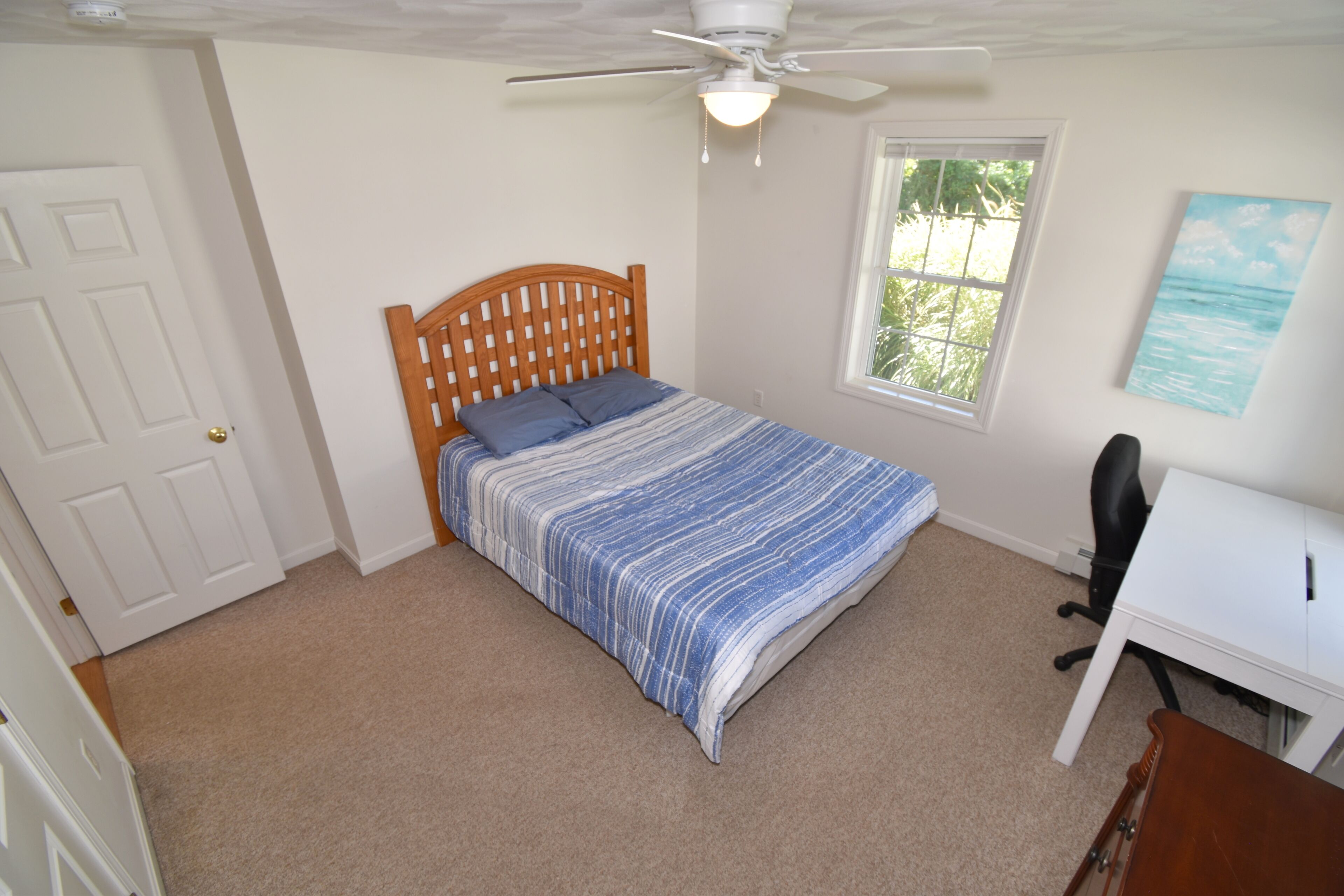 Ein Schlafzimmer mit Bett, Schreibtisch, Stuhl, Deckenventilator, Fenster und Tür.