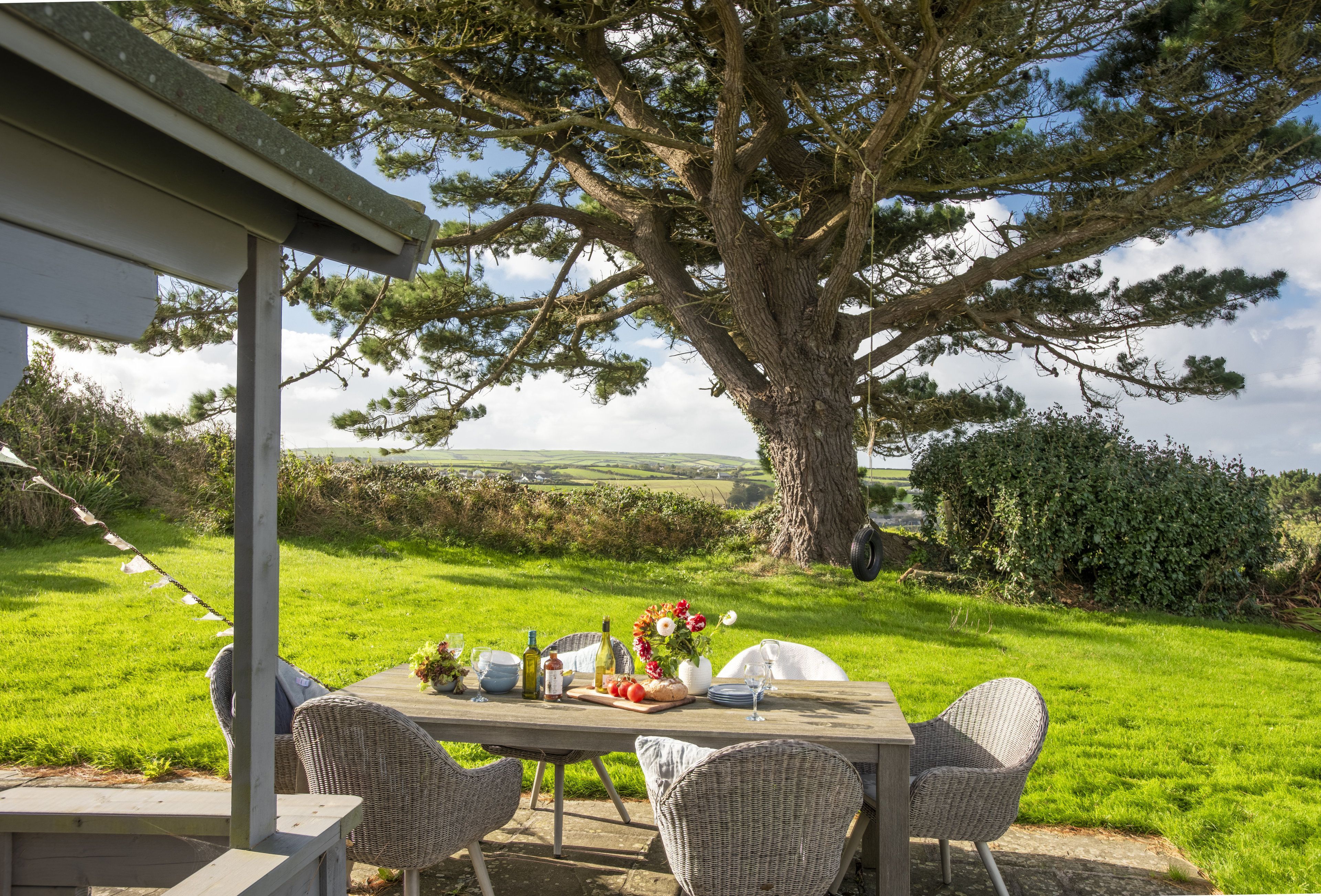 An outdoor dining area with a table set for two, surrounded by wicker chairs, under a large tree.