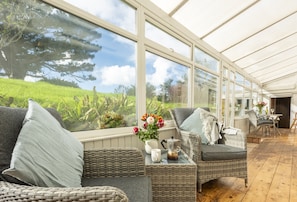 A conservatory with wicker furniture, a table with a kettle, and a view of a green landscape. - Little Penrose Cottage is a spacious and characterful home set in mature gardens (Bude)