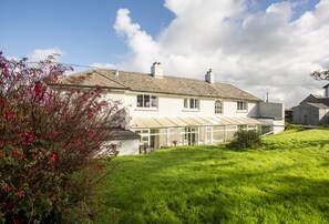 A two-story house with a large glass conservatory, a sloped roof, and a well-maintained lawn. - Little Penrose Cottage is a spacious and characterful home set in mature gardens (Bude)