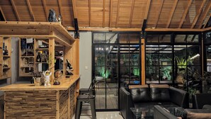 Un interior moderno con techo de madera, una zona de bar con estanterías, un área de estar con sofás de cuero y ventanas amplias con plantas.