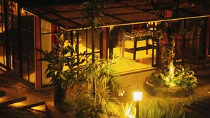 Vista nocturna de un edificio con fachada de vidrio, zona de estar al aire libre y plantas en macetas.