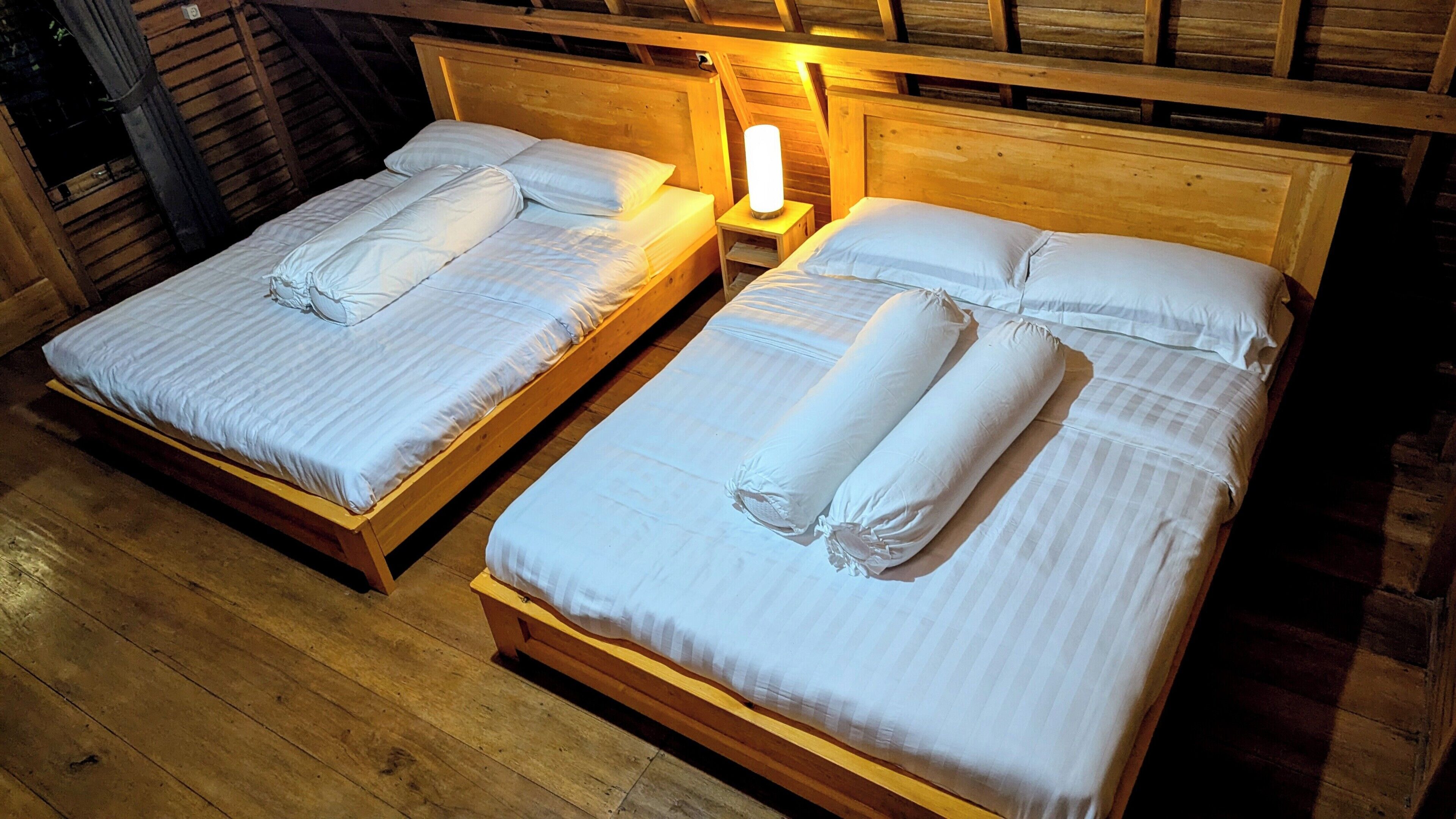 Dos camas de madera con sábanas y almohadas blancas, una mesita de noche con lámpara y paredes de madera.