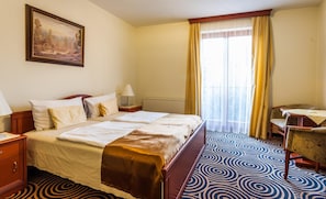 Habitación de hotel con cama doble, escritorio, silla, radiador, un cuadro y ventana con cortinas.