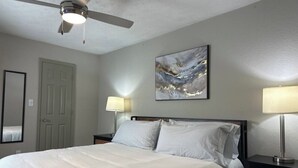 A hotel room with a bed, two bedside tables, a ceiling fan, a painting on the wall, and a door. - Lovely stay by the Braves / 2bd 2bth with private access gate to Braves Stadium (Smyrna)