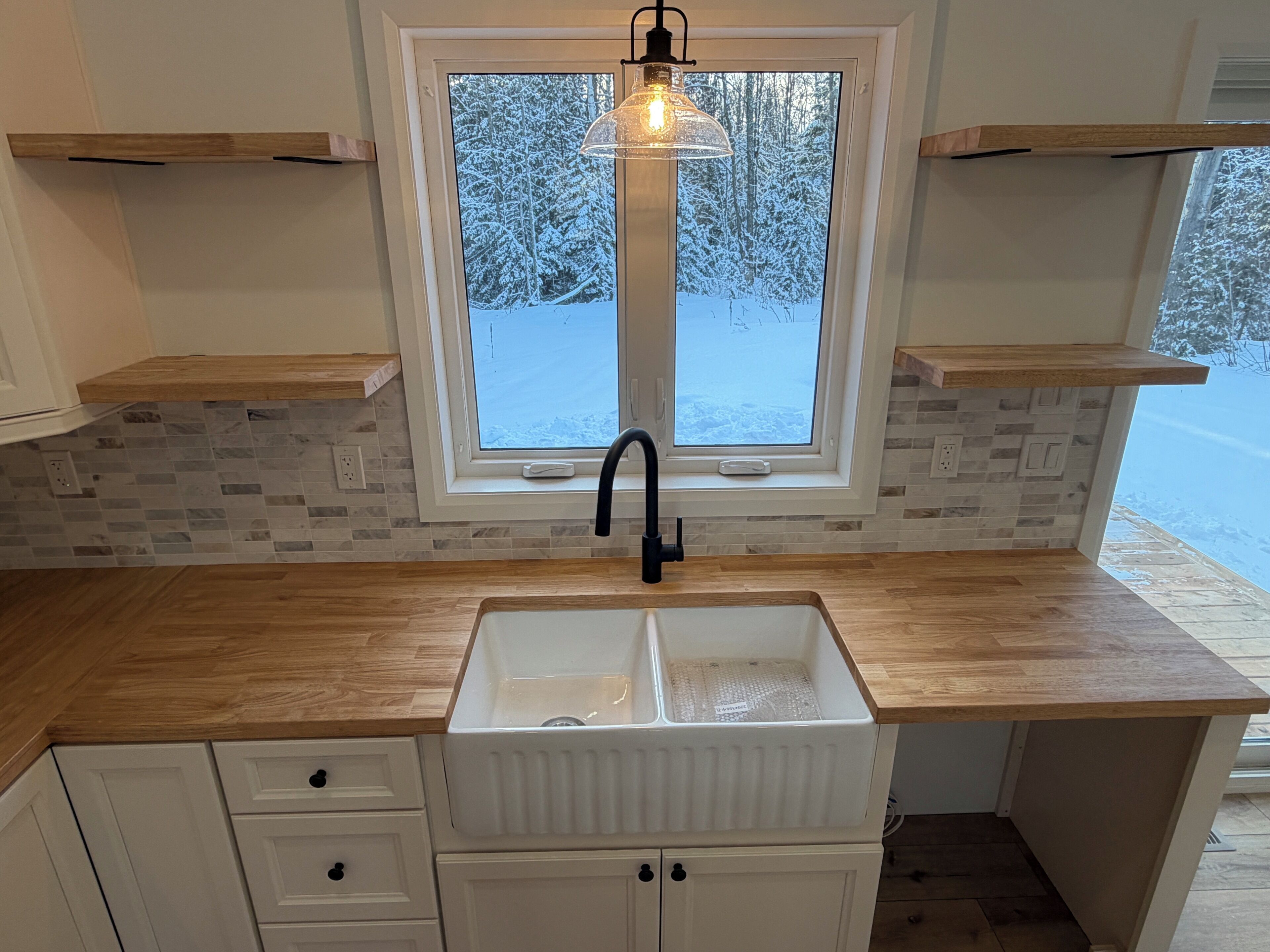 A kitchen with a double sink, wooden countertop, white cabinets, and a window with a view of a snowy landscape.