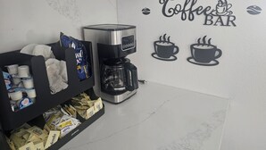 Een koffiehoek met een koffiezetapparaat, een plank met koffiecups en een muursticker met de tekst 'Coffee BAR'.