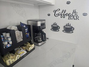 Ein Kaffee-Bereich mit Kaffeemaschine, einer Ablage für Kaffeekapseln und einer Wanddekoration mit der Aufschrift 'Coffee BAR'.