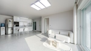 A modern kitchen with white cabinetry, a dining area with a white table and chairs, a white sofa, and a large window with natural light. - Yeongheungdo Jeongwonstay (Incheon)