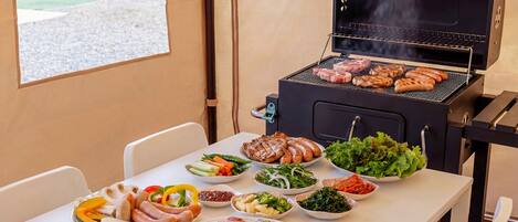 Un'area barbecue con diversi tipi di carne e verdure su piatti, una griglia e un tavolo con sedie.