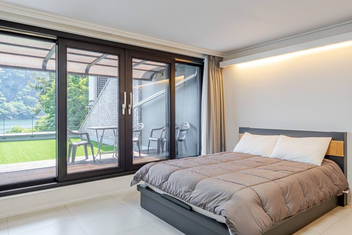 Un dormitorio con cama, puerta corrediza al balcón y vistas a un edificio y follaje.