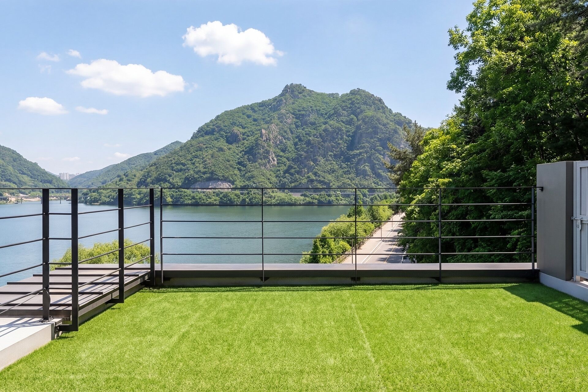 A terrace with a view of a lake and mountains, a metal railing, and a paved walkway.