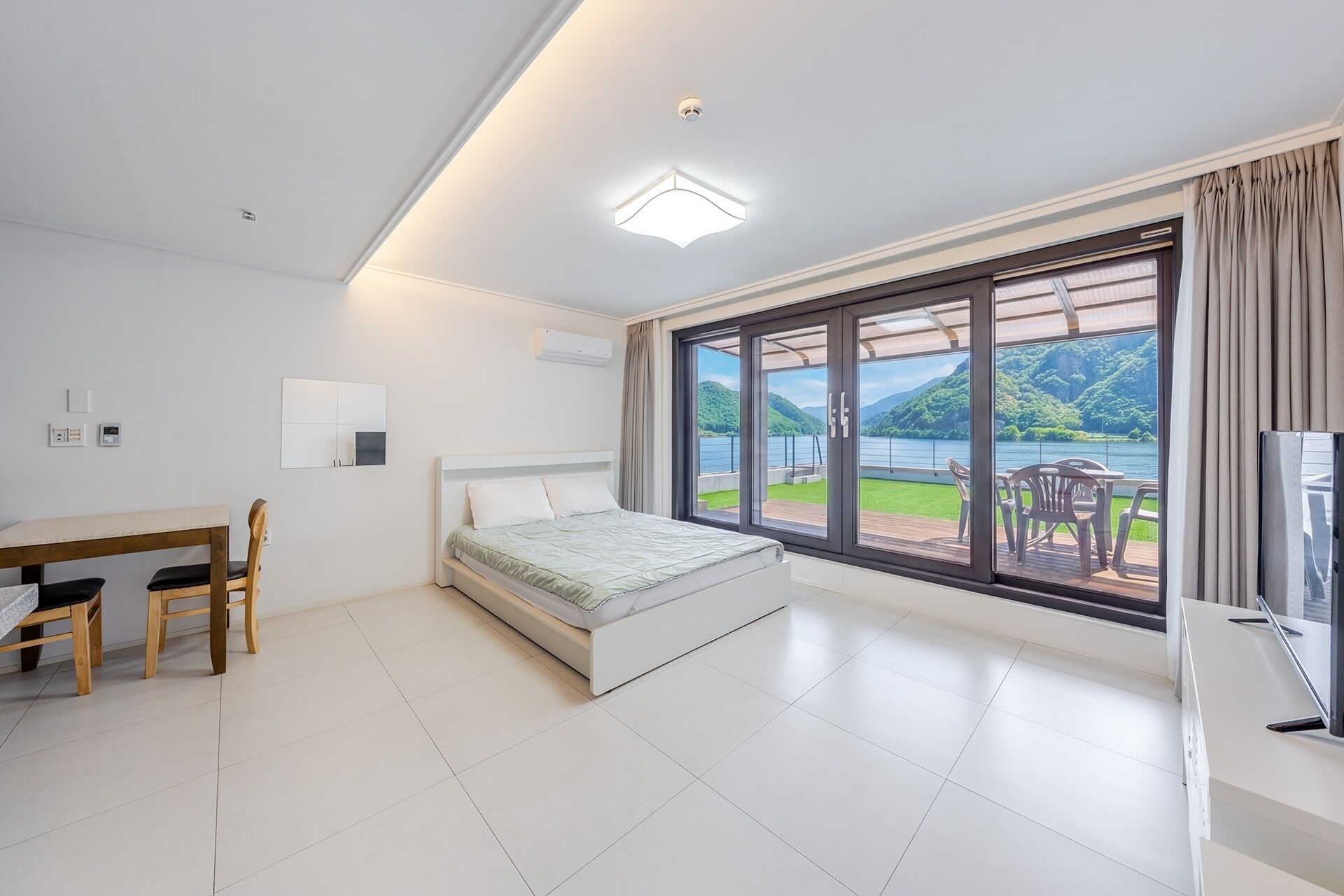 A bedroom with a bed, a desk, chairs, a TV, and a large sliding glass door leading to a balcony with a view of a lake and hills.