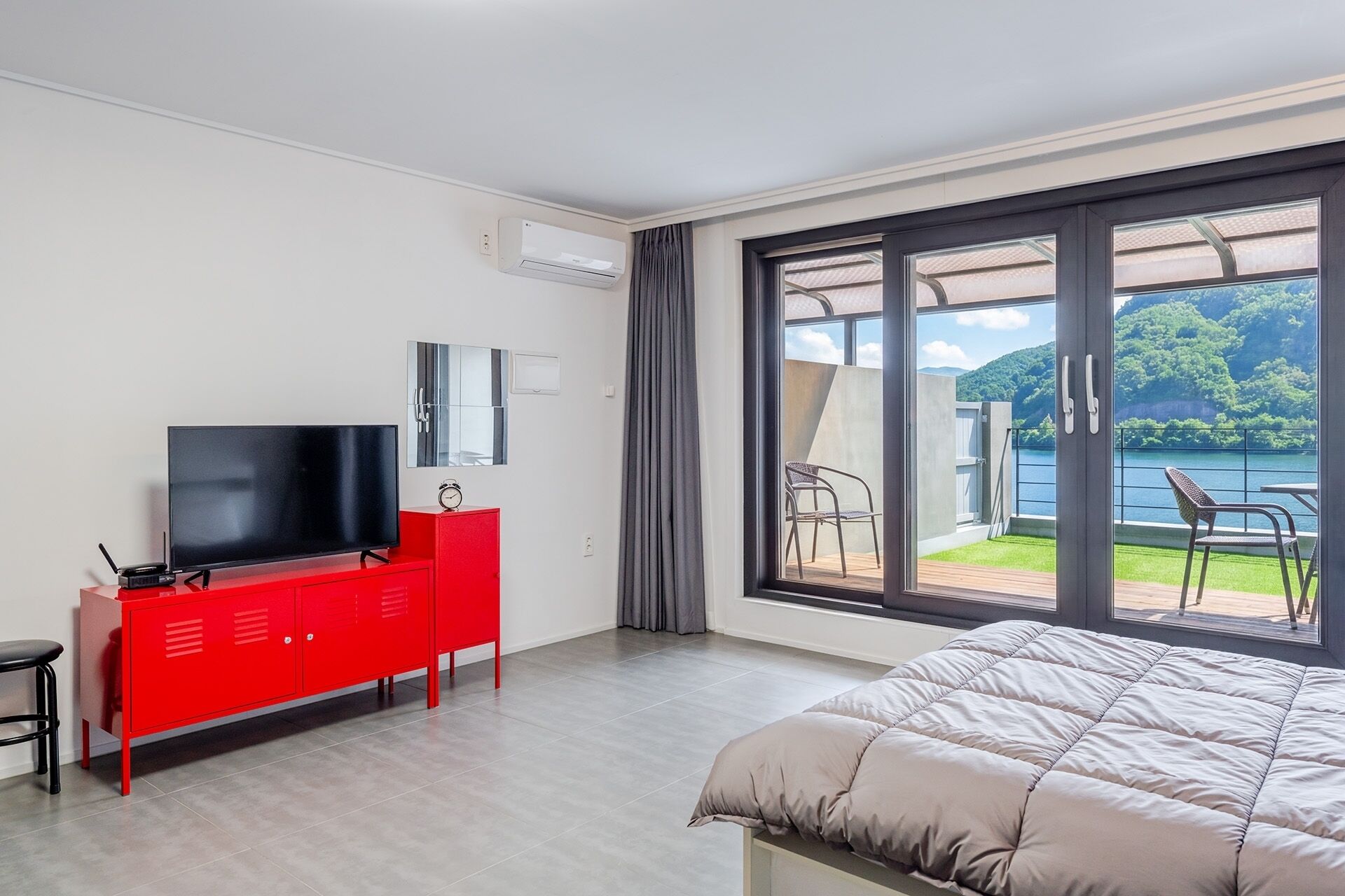 A hotel room with a bed, a red dresser with a TV, a window with a view of a lake and hills, and a balcony with chairs.