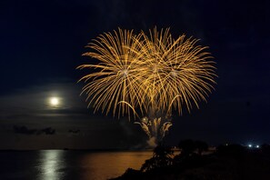 A night scene with fireworks over a body of water, a visible moon, and silhouettes of trees and distant lights. - Uruma Dome Okinawa Saturn7 (Uruma)