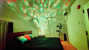 A bedroom with a bed, a nightstand, a lamp, a plant, and a projection of colorful lights on the ceiling. - Uruma Dome Okinawa Saturn7 (Uruma)