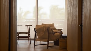 Una habitación con piso de madera, una silla de madera, un sillón con cojín y una manta, y una mesita.