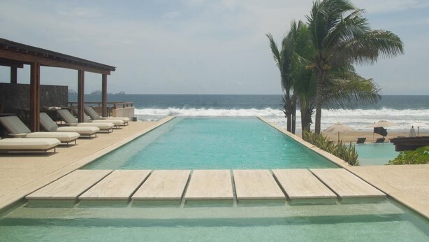 Una piscina con sillones de descanso, una pérgola de madera, palmeras y vista al océano.