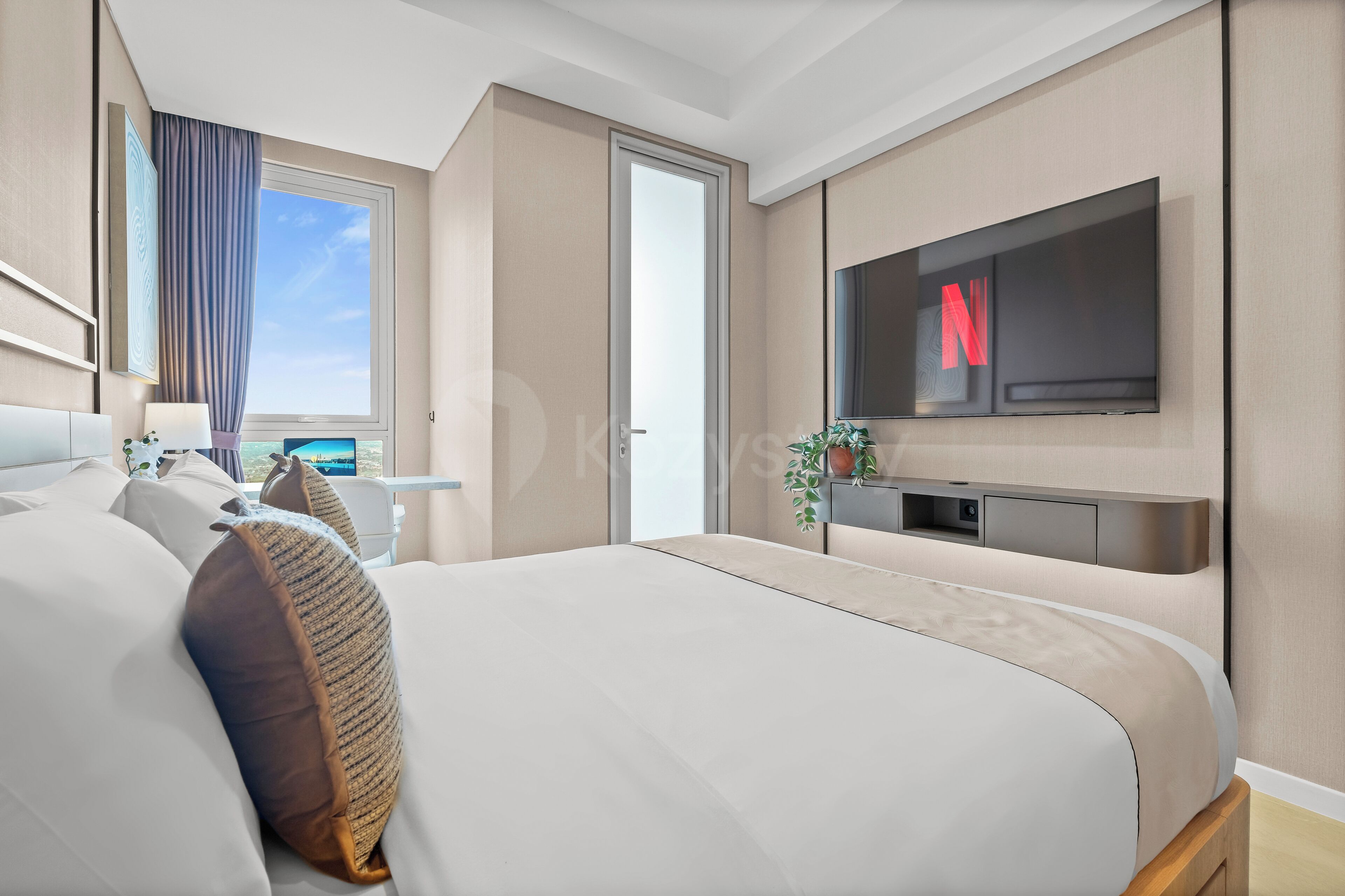 Une chambre d’hôtel moderne avec un grand lit, un téléviseur fixé au mur, un bureau avec une lampe et une vue sur le ciel par une fenêtre.