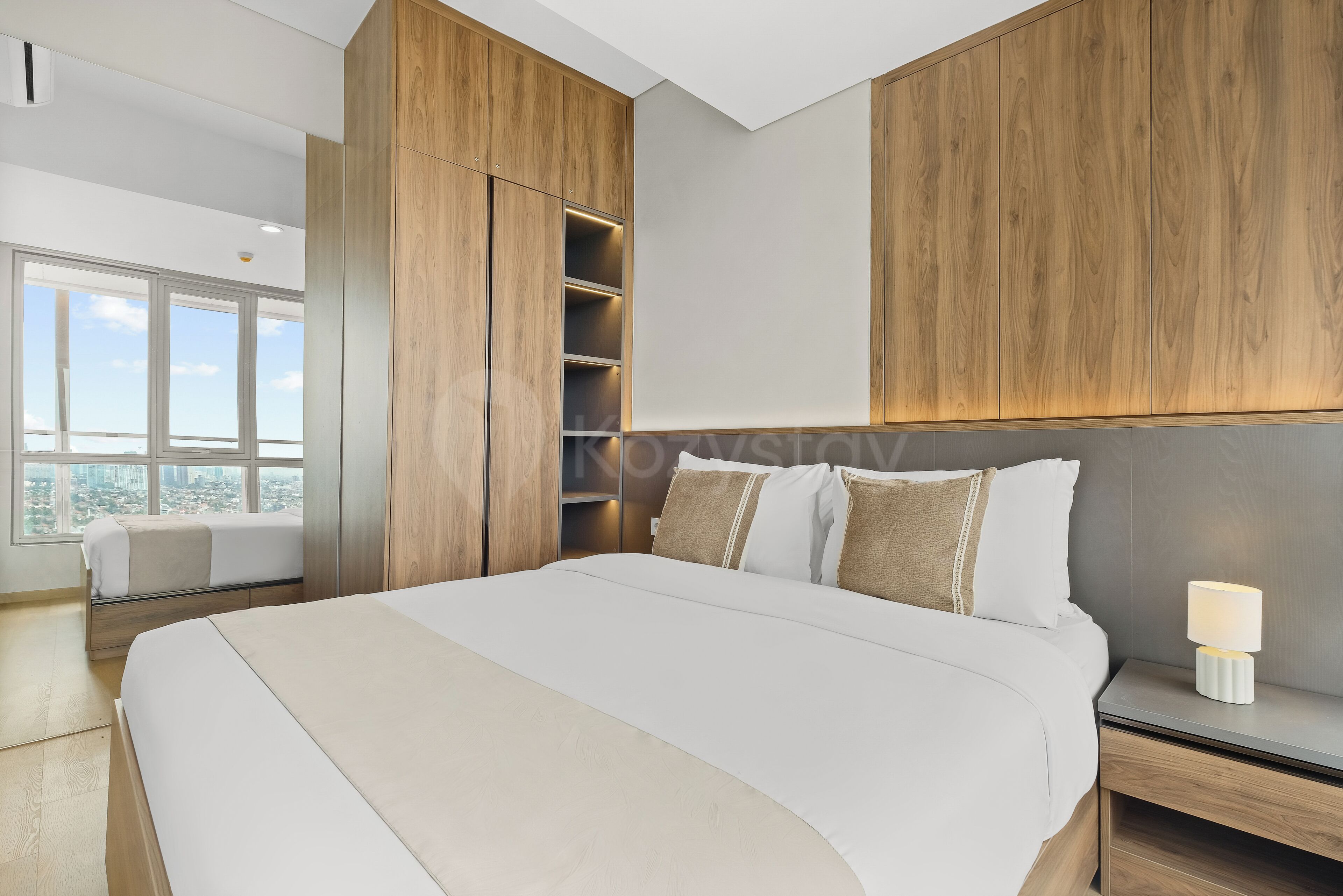 A modern bedroom with a large bed, built-in wooden shelving, a bedside table with a lamp, and a view of the city through large windows.