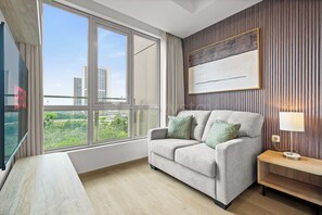 A modern hotel room with a large window, a flat-screen TV, a sofa with cushions, a side table with a lamp, and a framed artwork on the wall. - Premium Apartment by Kozystay - Antasari Place (Kota Jakarta Selatan)