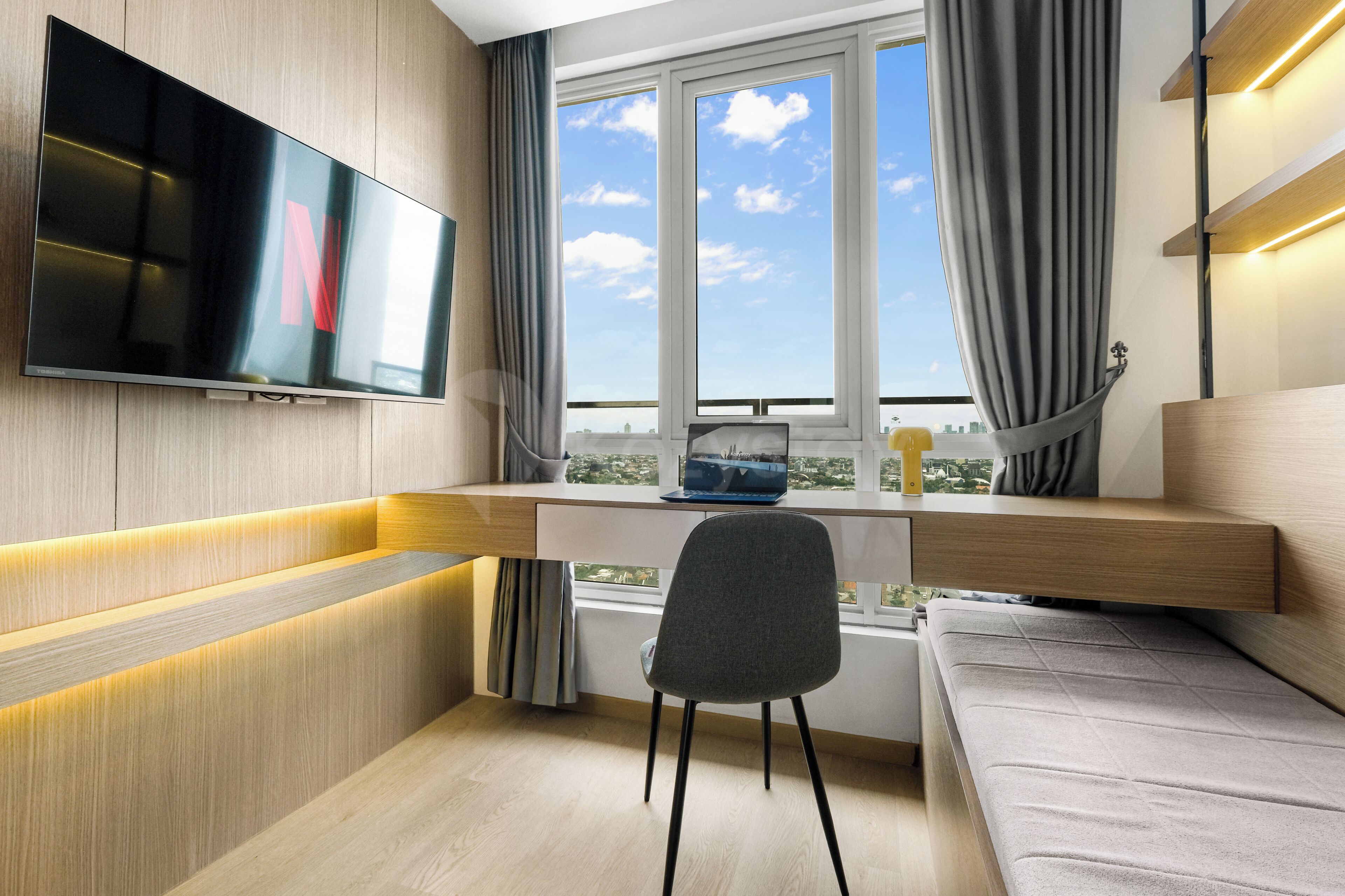 A modern room with a flat-screen TV mounted on the wall, a desk with a laptop and a cup, a grey chair, and a sofa.
