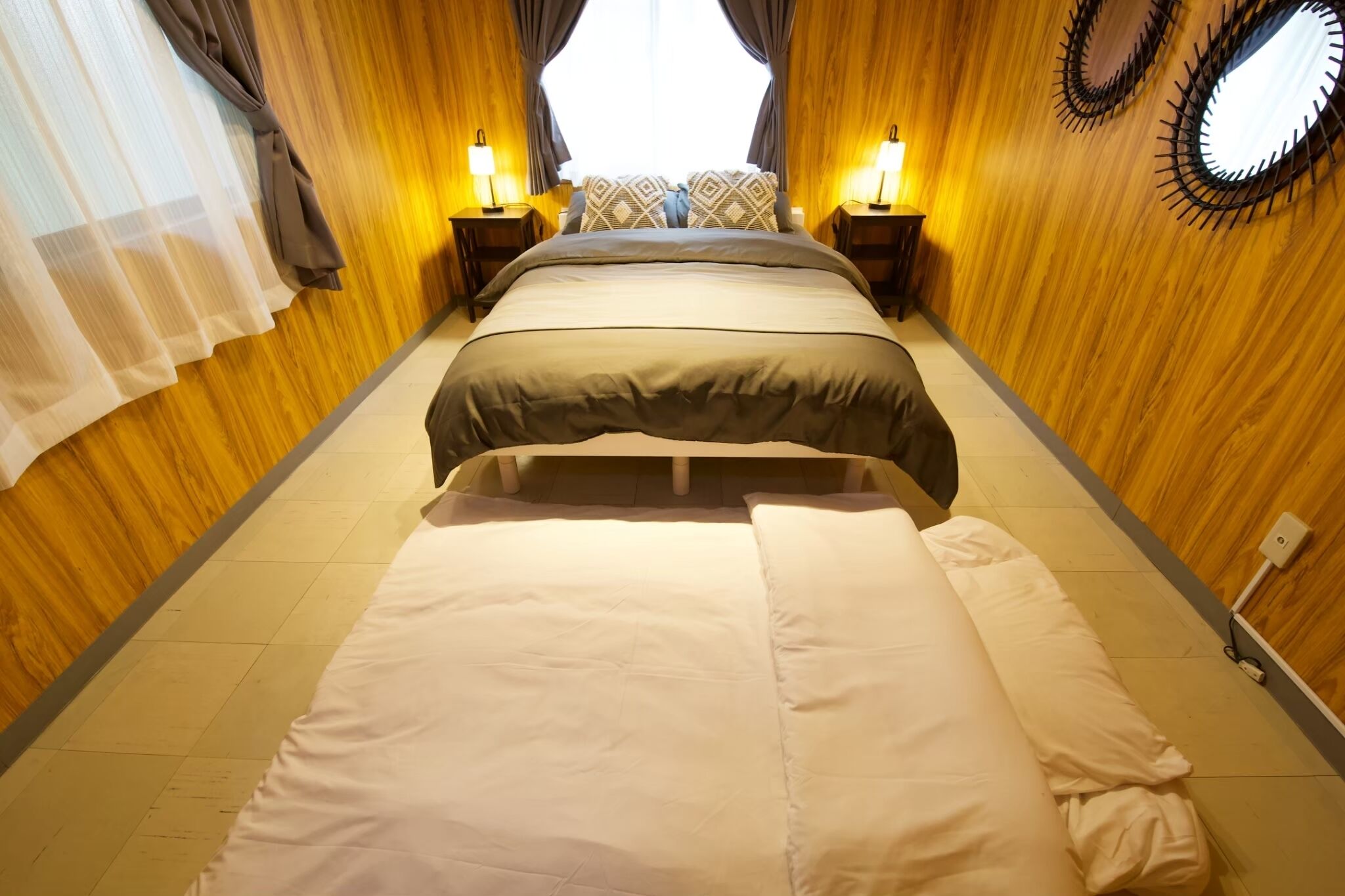 A hotel room with a bed, bedside lamps, a window with curtains, and a wooden wall.