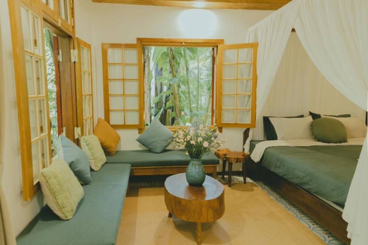 A room with a bed, a sofa, a round wooden table, and a window with a view of greenery.