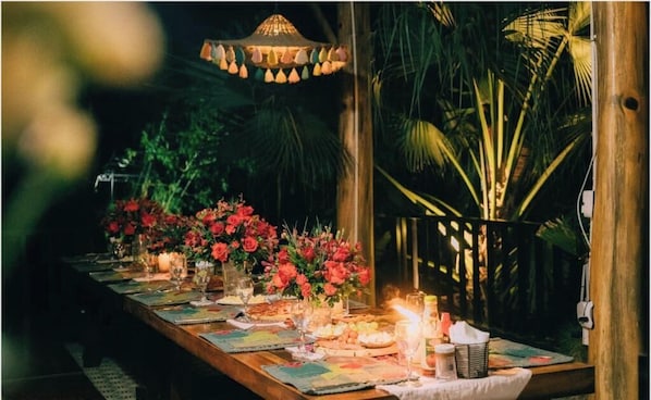 Un área de comedor al aire libre con una mesa de madera dispuesta para una comida, rodeada de abundante follaje y una lámpara colgante decorativa.