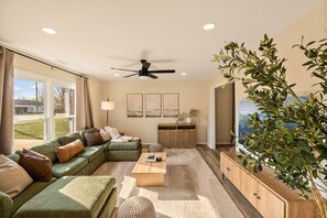 Una sala de estar con un sofá modular, una mesa de centro, un ventilador de techo, un televisor y una planta grande.