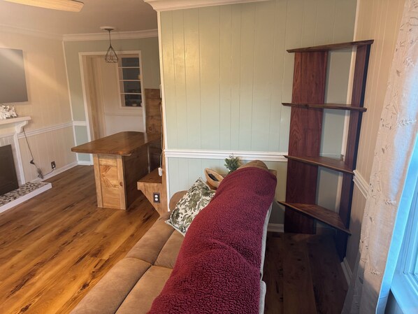 Una sala con suelo de madera, un sofá, una manta roja, un mueble de madera y una puerta que da a otra habitación.