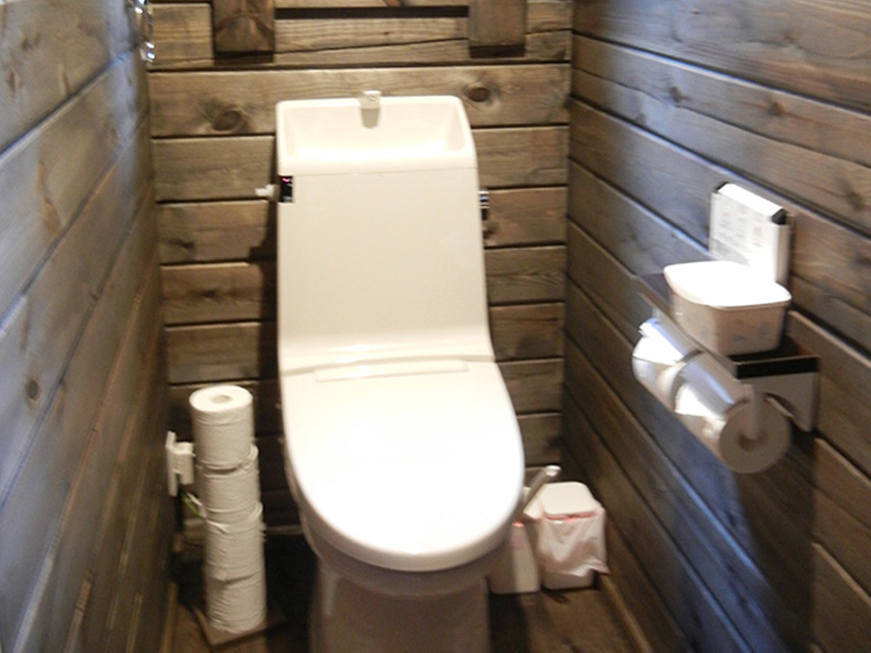 A small, narrow bathroom with wooden walls, a white toilet, and a toilet paper holder.