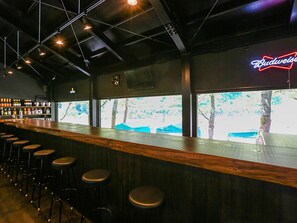 A bar area with wooden stools, a neon Budweiser sign, and a view of trees through large windows. - Nagomiya Seseragi (Gujo)