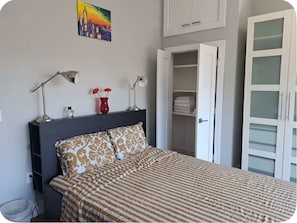 A bedroom with a bed, bedside lamps, a red vase with flowers, a cityscape painting, a wardrobe, and a closet with folded clothes.