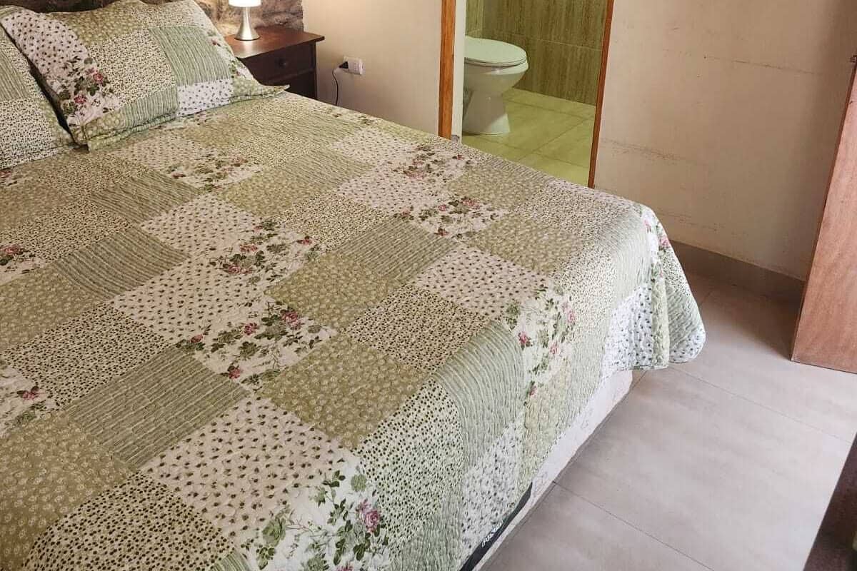 Un dormitorio con pared de piedra, una cama con colcha estampada, una mesita de noche con lámpara y un baño visible a través de una puerta abierta.