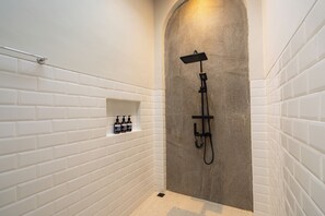 A bathroom with white tiled walls, a shower area with a stone accent wall, and a shelf with toiletries. - Serenia Villa by Betterplace (Ubud)