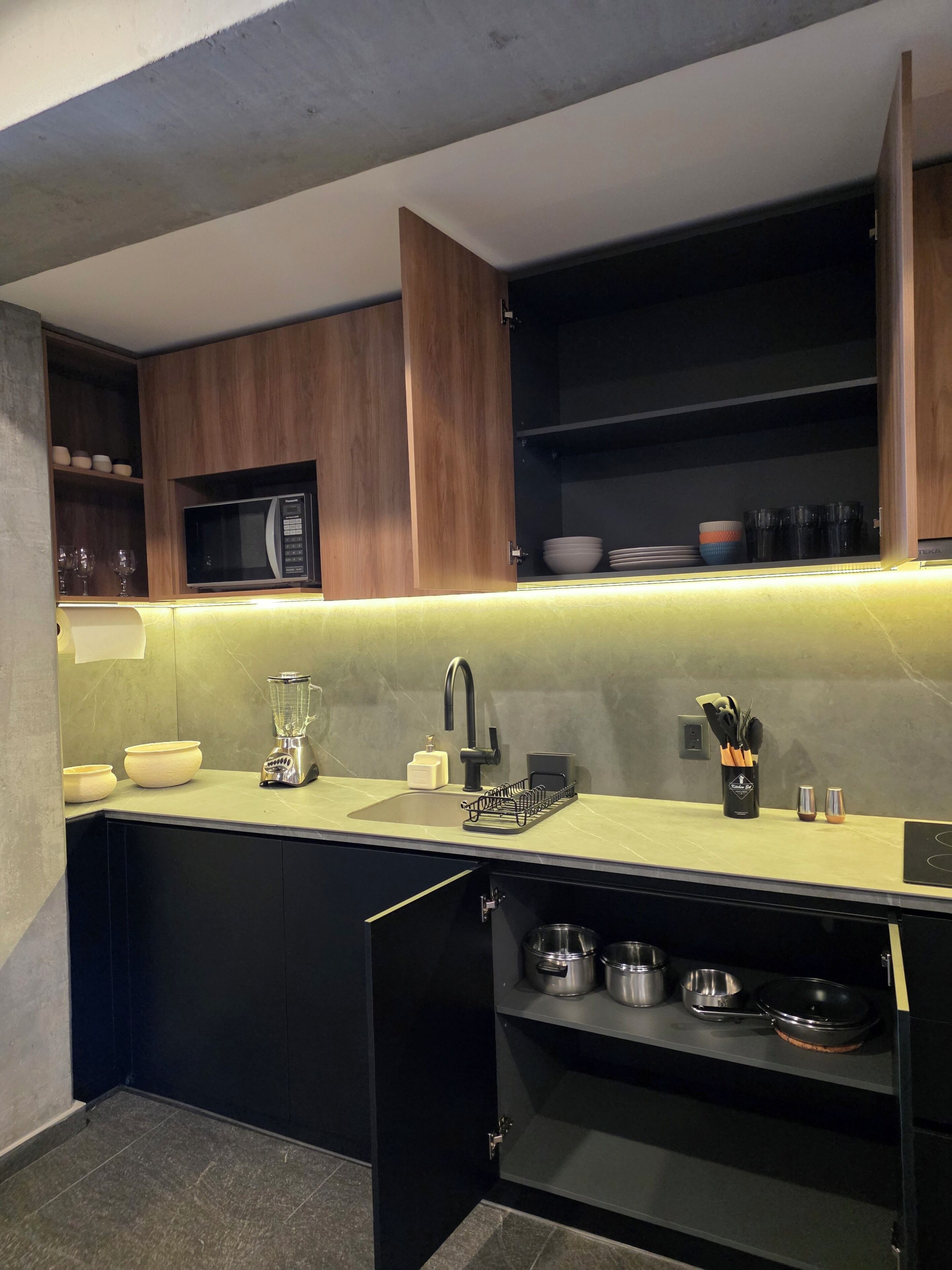 Una cucina moderna con forno a microonde integrato, lavello con rubinetto, frullatore e scolapiatti. Sono presenti pensili sopra il piano di lavoro e un ripiano inferiore con pentole e padelle.